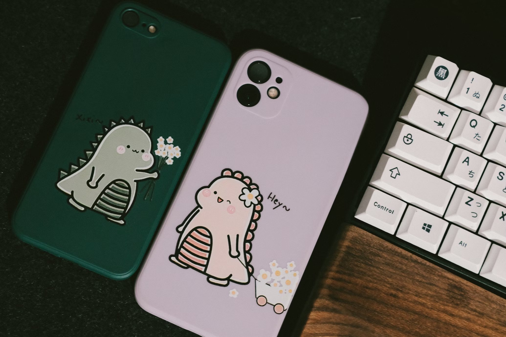 Capas em dois smartphones, próximos de um teclado. As capas possuem dois dinossauros fofinhos a darem flores um ao outro