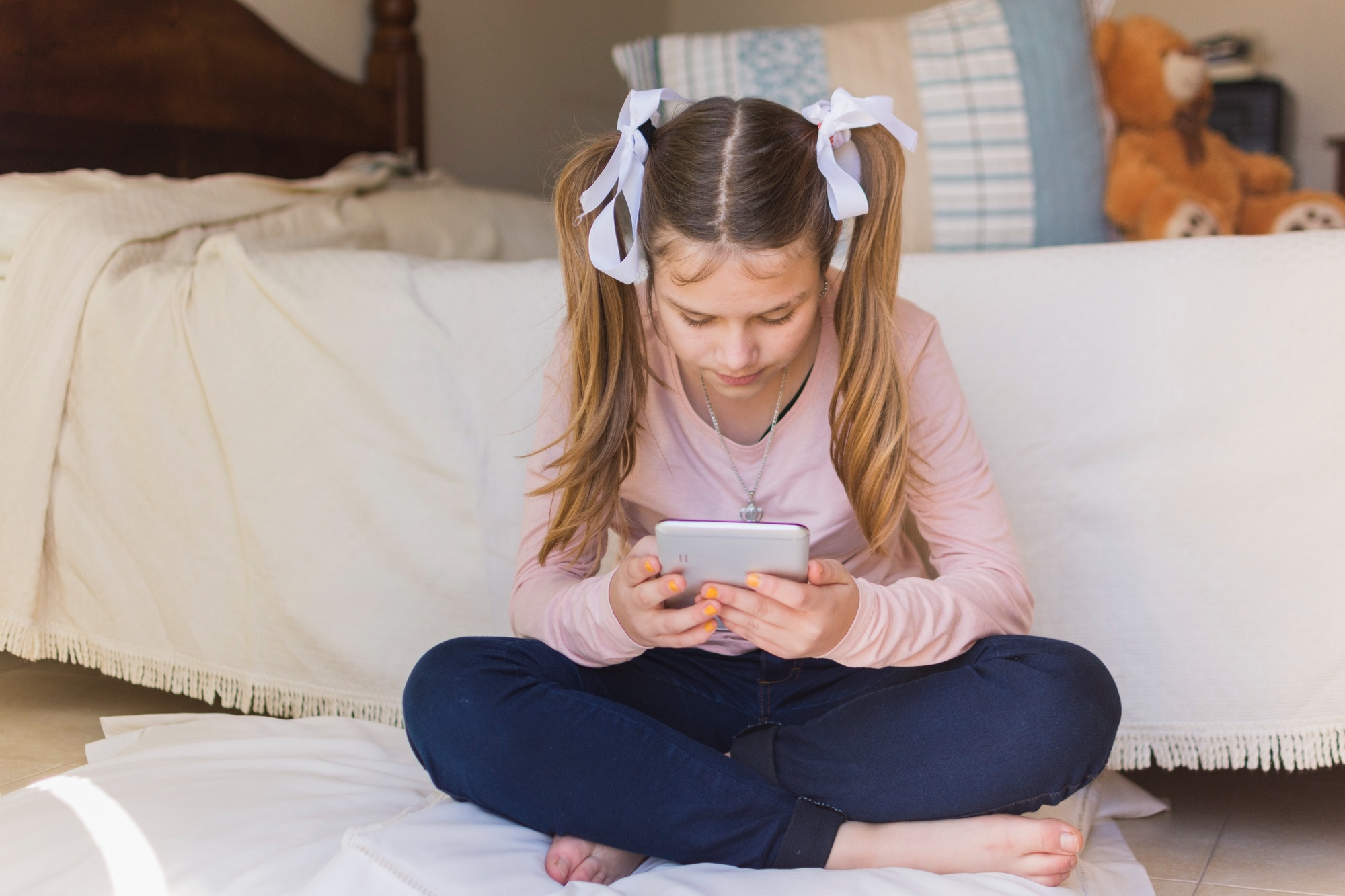 criança a usar smartphone, sentada no chão, em frente de uma cama
